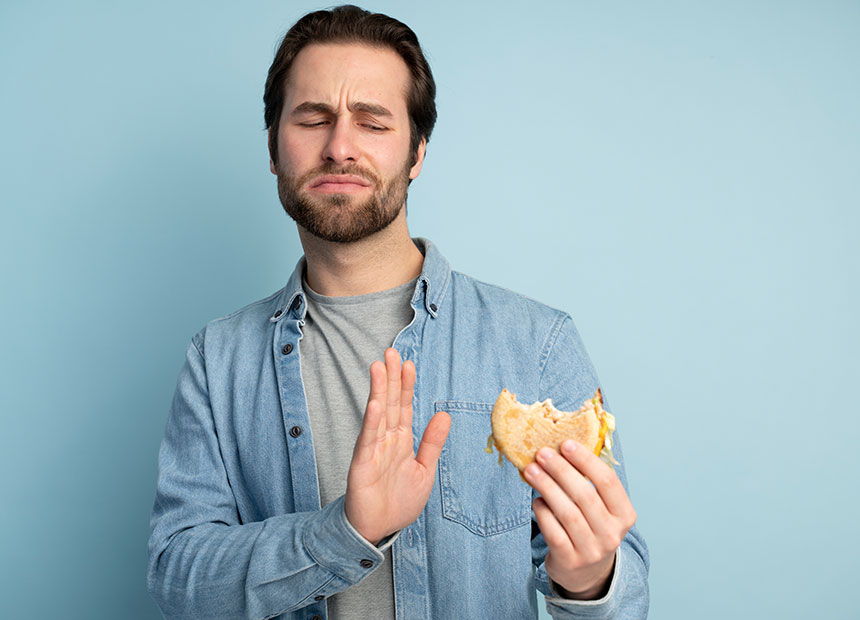 Man in a denim shirt holding a half-eaten sandwich in one hand and gesturing stop with the other hand against a light blue background.